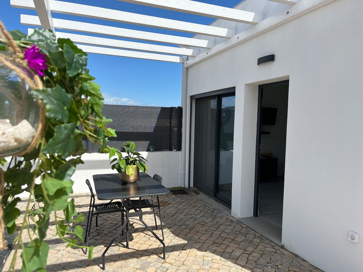 An outdoor patio area is featured, showcasing a black dining table surrounded by four chairs. Green foliage hangs above, and sunlight brightens the space. The sliding glass door leads to an interior area, while a clear blue sky is visible in the background.