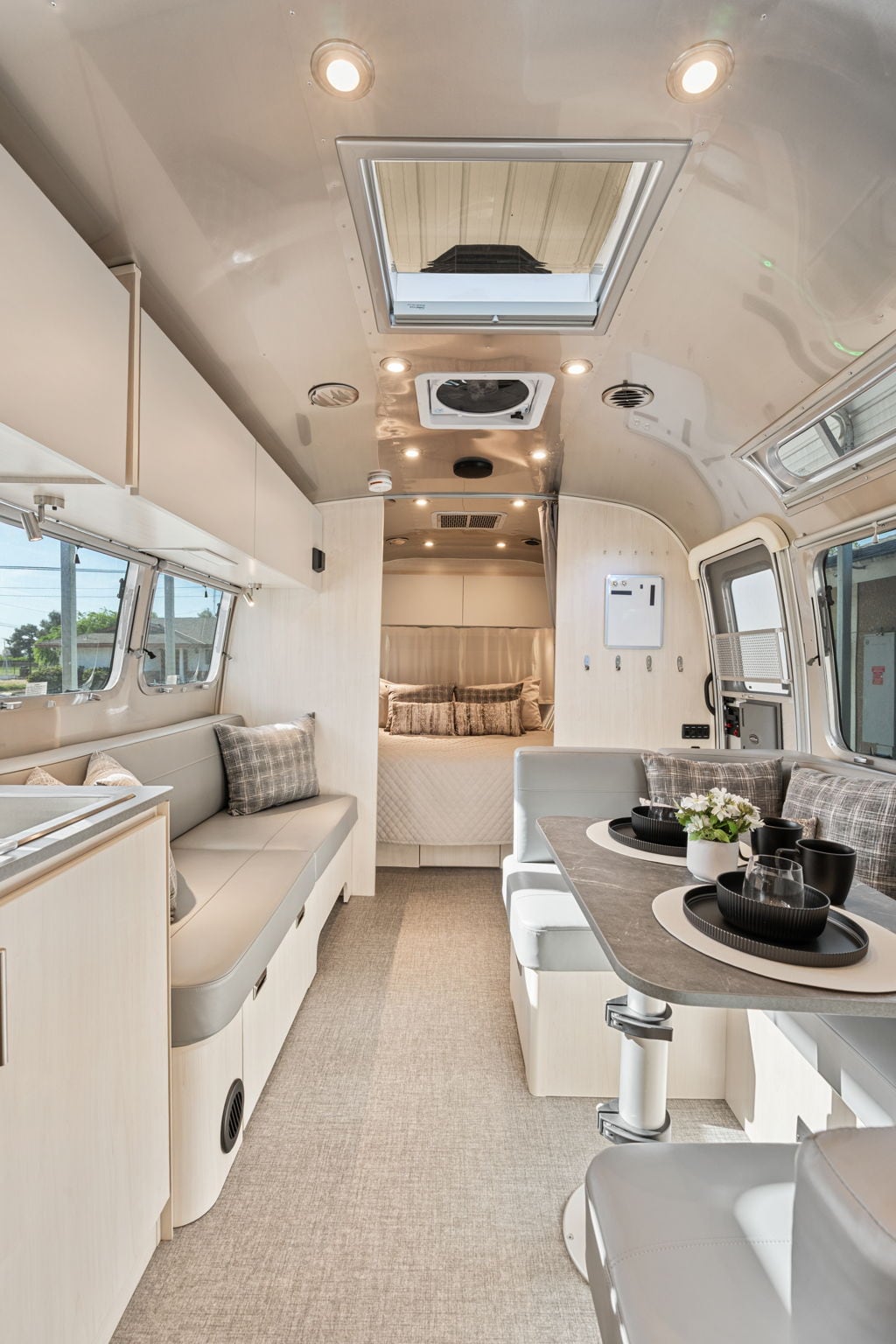 The interior of the Airstream is illuminated by soft overhead lighting, featuring a cozy seating area with plush cushions. A dining table is set with dishes and utensils, while the spacious layout leads to a sleeping area in the background, separated by a subtle divider.