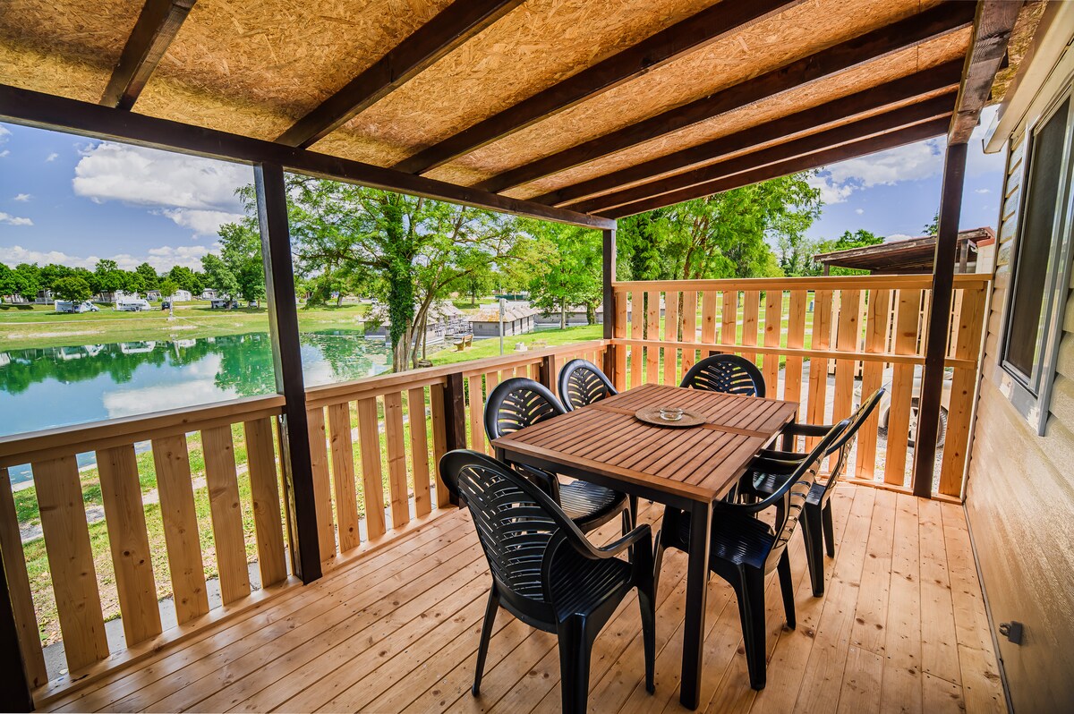 An outdoor dining area features a wooden table surrounded by six black chairs, offering a scenic view of the nearby water and greenery. The space is partially covered by a wooden roof, providing shade and a relaxing atmosphere.