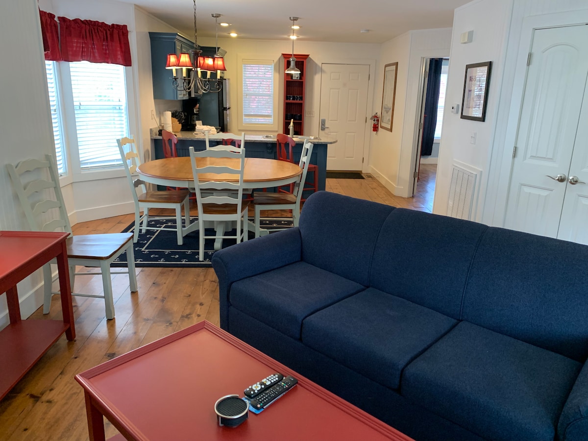 A cozy living area features a navy blue sofa facing a dining table with circular design. Red accent tables are positioned nearby, and a kitchen area is visible in the background. Light enters through multiple windows, enhancing the space's brightness.