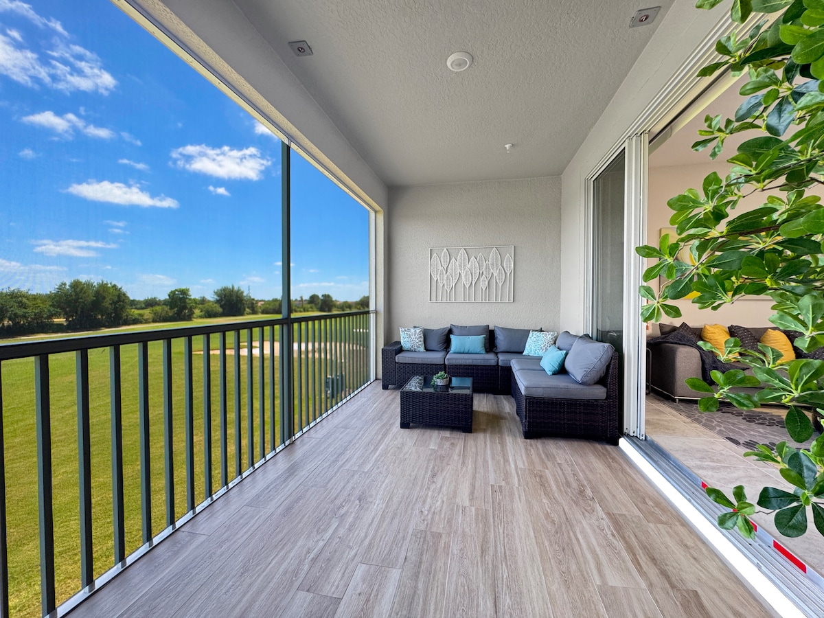 A spacious balcony overlooks lush green golf course views, featuring a comfortable seating area with dark wicker furniture and light blue cushions. Large sliding glass doors provide easy access to the interior space, while sunlight filters through the open layout.