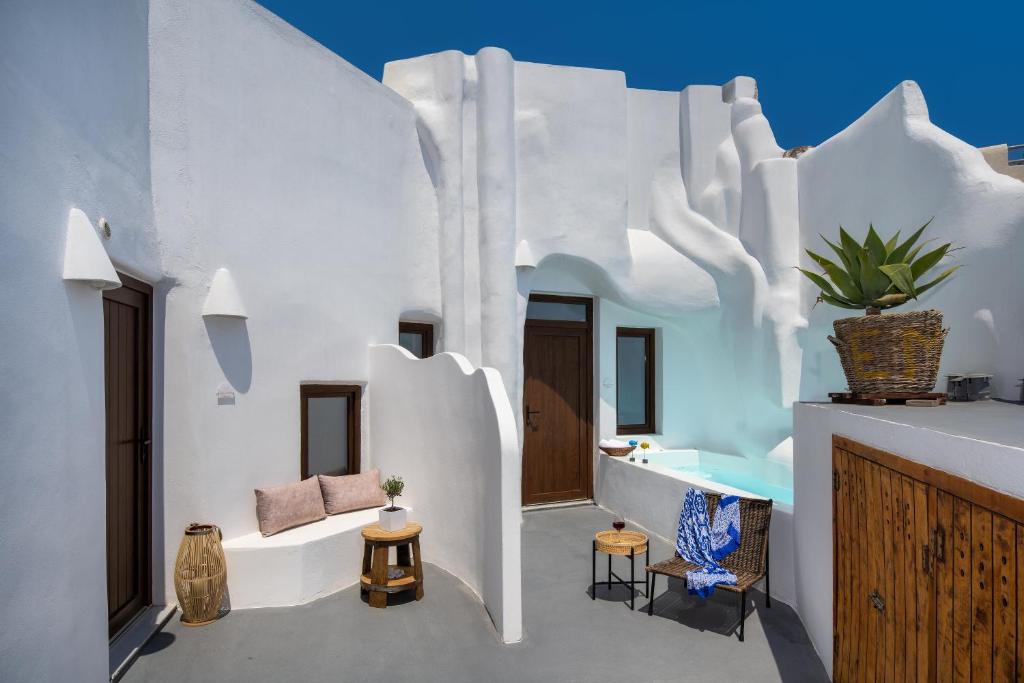 An outdoor area is shown, featuring whitewashed walls and sculptural architectural details. A small swimming pool can be seen beside a plant in a woven basket. A seating area with a small table, woven chair, and decorative elements is located nearby, providing a relaxing space.