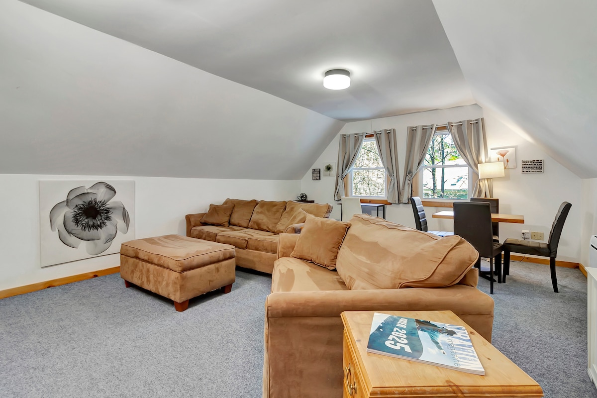 A spacious living area features a large sectional sofa and an ottoman, complemented by a dining table with chairs. Natural light filters through the windows adorned with light curtains, and a floral artwork adds a touch of character on one wall.