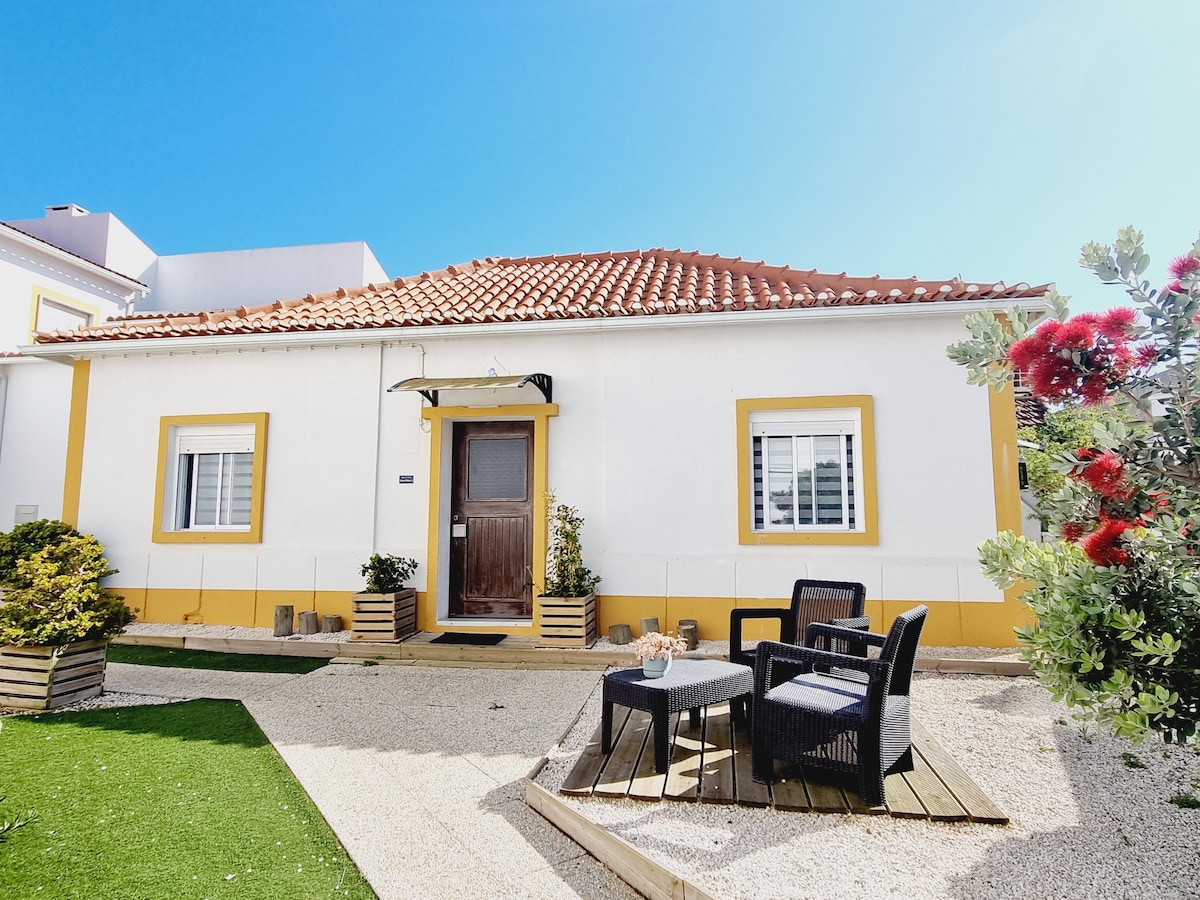 The exterior of a renovated home is highlighted, showcasing a welcoming entrance with a wooden door. The space is complemented by two outdoor chairs and a small table, surrounded by well-maintained greenery and a gravel pathway under clear blue skies.