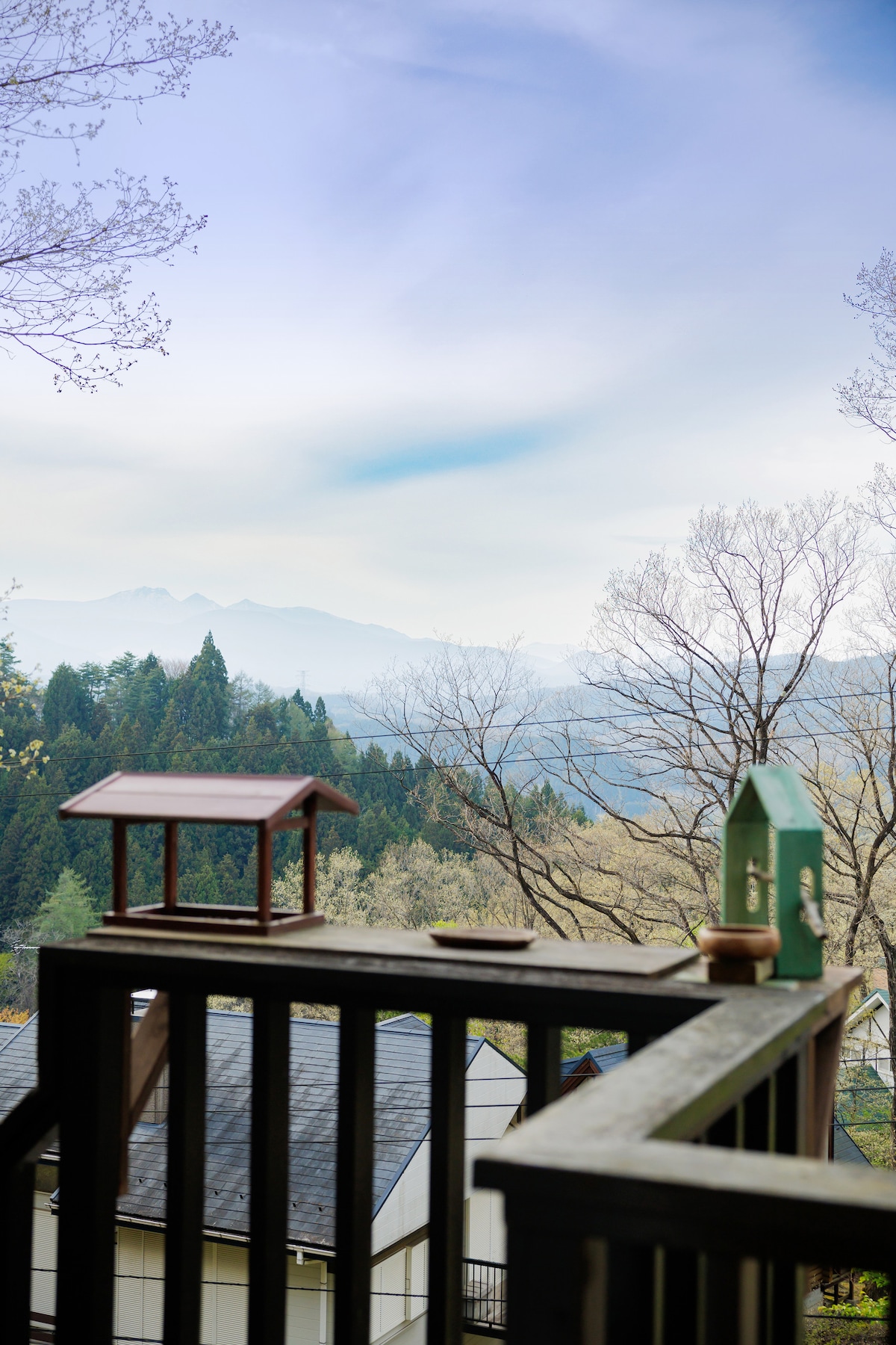 A wooden deck railing frames a scenic view of the mountains, partially shrouded in mist. A birdhouse is positioned nearby, inviting wildlife. Leafless trees extend in the background, creating a serene connection with nature. Soft blue skies complement the tranquil landscape.