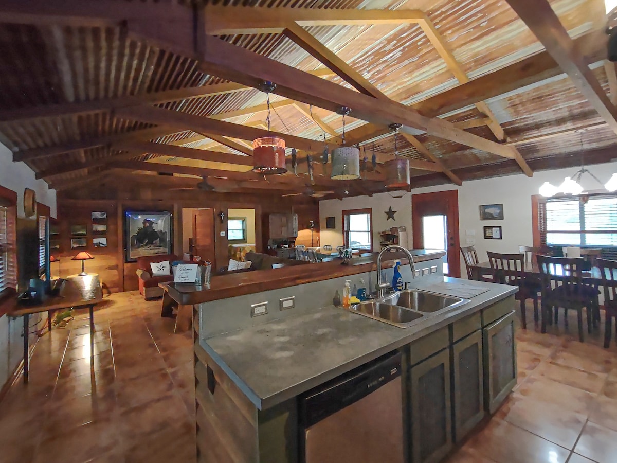 The kitchen features an open layout with wooden beams overhead and rustic decor. Stainless steel appliances are visible, along with a central island and sink. The living area is seen in the background, showcasing comfortable seating and natural light from nearby windows.