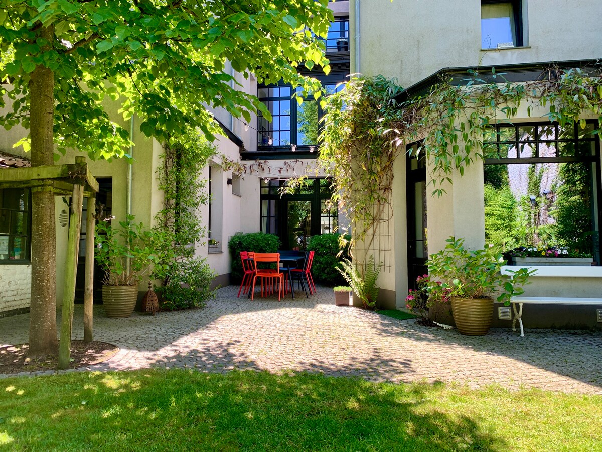 A sunny garden area features a stone-paved surface with a set of bright red outdoor chairs and a table. Lush greenery surrounds the space, with trees and potted plants adding to the inviting atmosphere. Large windows from the house overlook the garden.