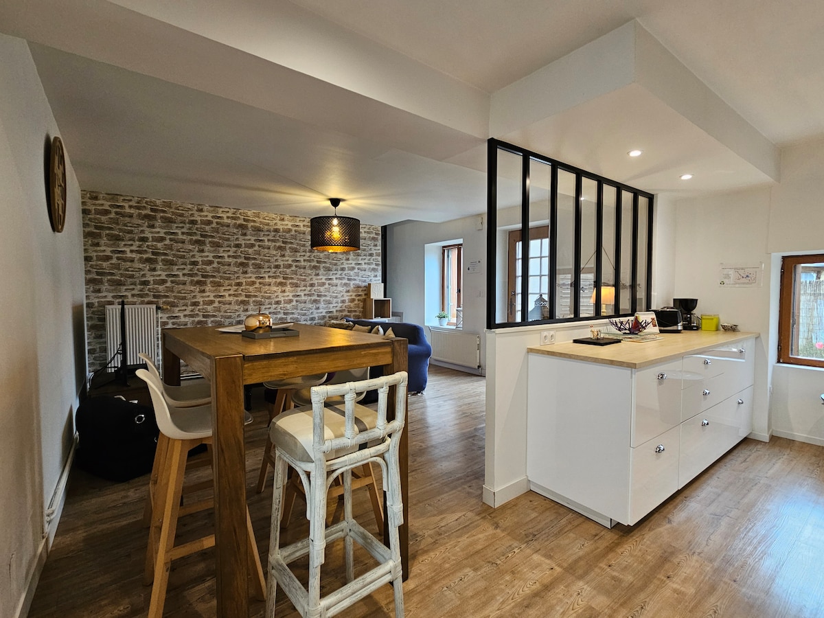 An open-plan kitchen and dining area features a wooden table accompanied by four stools. Light-colored cabinetry offers ample storage. A wall of exposed stone adds texture, while large windows provide natural light. A cozy seating area is visible in the background.