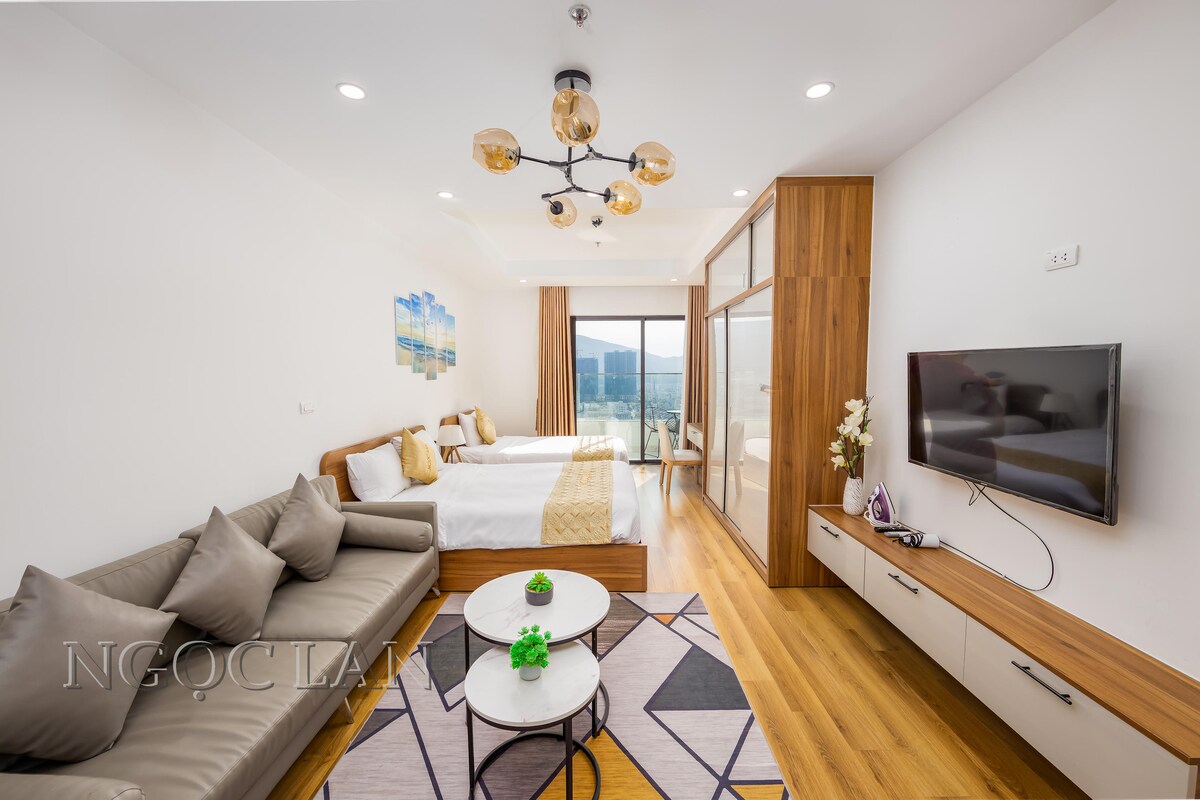 A spacious living area features a comfortable sofa with decorative pillows, accompanied by a coffee table. Two beds are positioned against a wall with a light wood accent. A flat-screen TV is mounted, and floor-to-ceiling windows provide an abundance of natural light and city views.