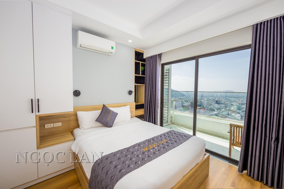 A comfortable bedroom features a spacious bed with soft bedding and a decorative throw. Large windows offer unobstructed views of the city and sea, while soft curtains frame the scene. A minimalist wardrobe and air conditioning unit are included in the well-designed space.