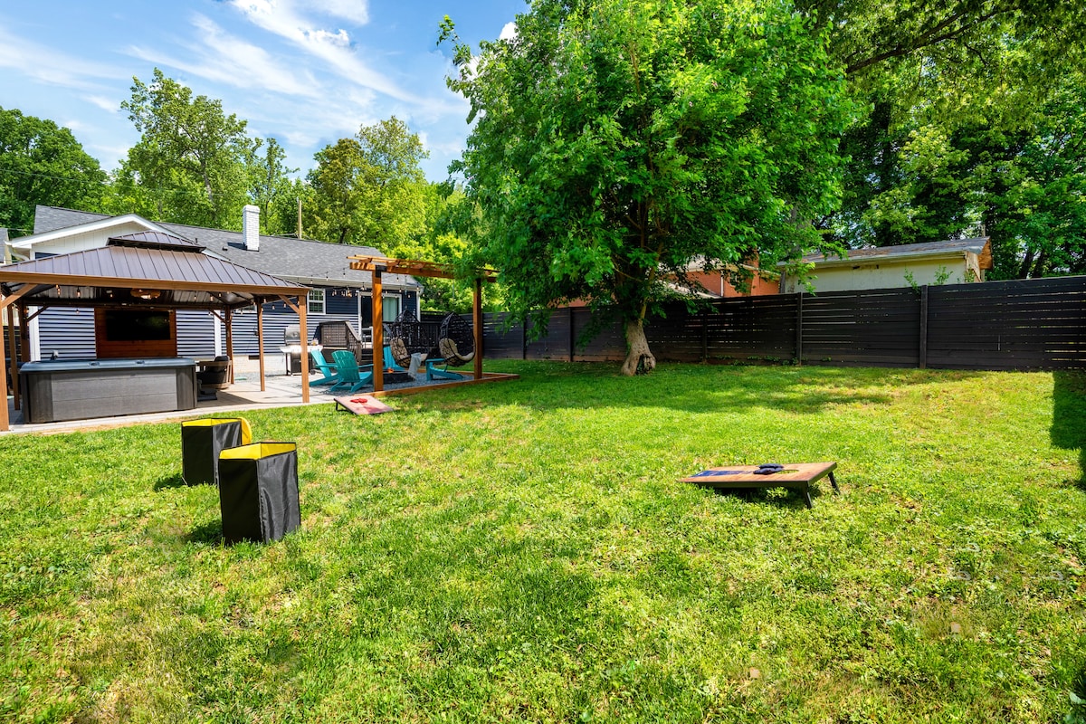 A spacious backyard is shown, featuring a gazebo with swing chairs and a hardscape area with a bar and outdoor TV. The lush green lawn provides ample room for yard games. The surrounding trees add natural shade to the inviting outdoor space.