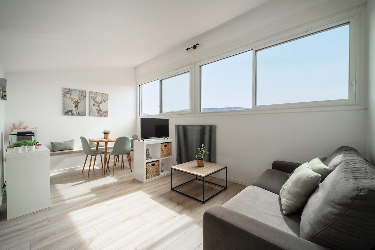 A bright living space is showcased, featuring large windows that offer views of the surrounding landscape. A comfortable gray sofa is positioned next to a coffee table, while a dining area with a round table and chairs is visible in the background. Neutral tones are used throughout.
