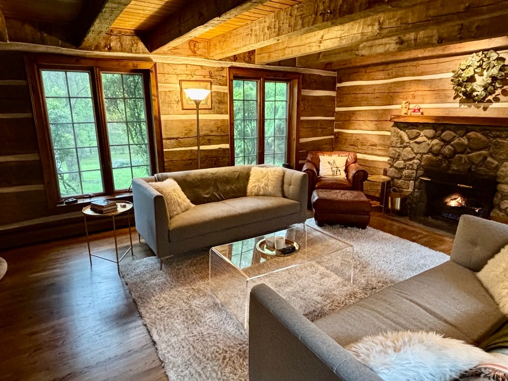A cozy living area features a stone fireplace, with comfortable seating on a plush rug. Large windows provide views of the surrounding greenery, while the warm tones of the wooden walls enhance the cabin's rustic charm. A floor lamp offers additional light.