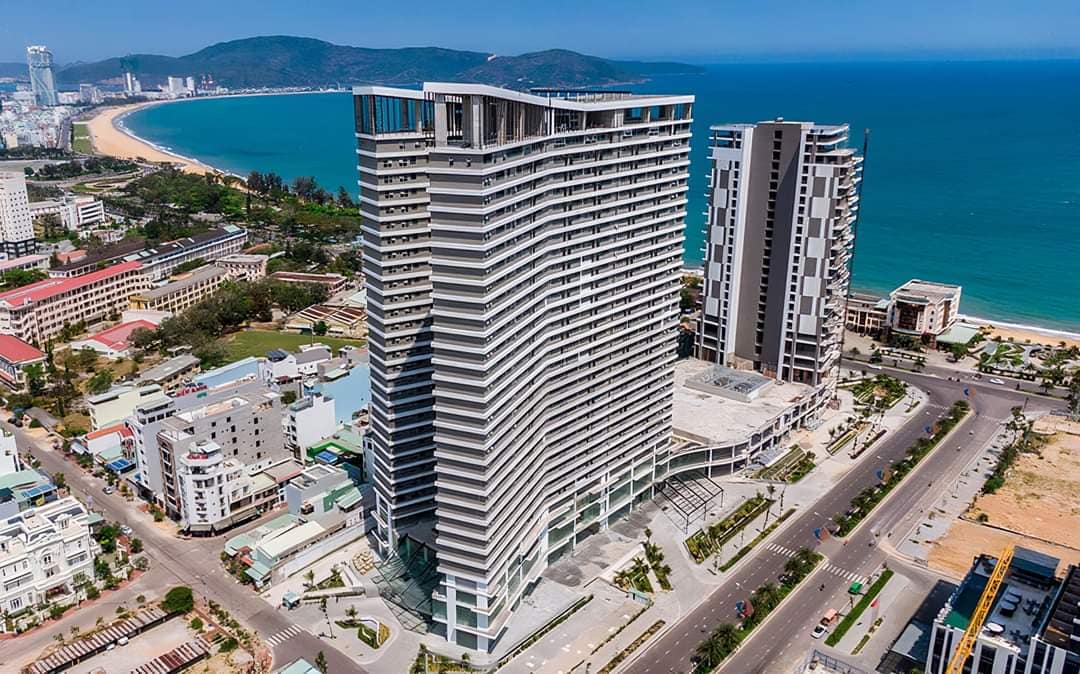 A modern high-rise building is prominently featured, overlooking the sea. The structure showcases a unique architectural design with curved balconies, while the shoreline and expansive ocean are visible in the background, along with stretches of sandy beach and nearby urban structures.