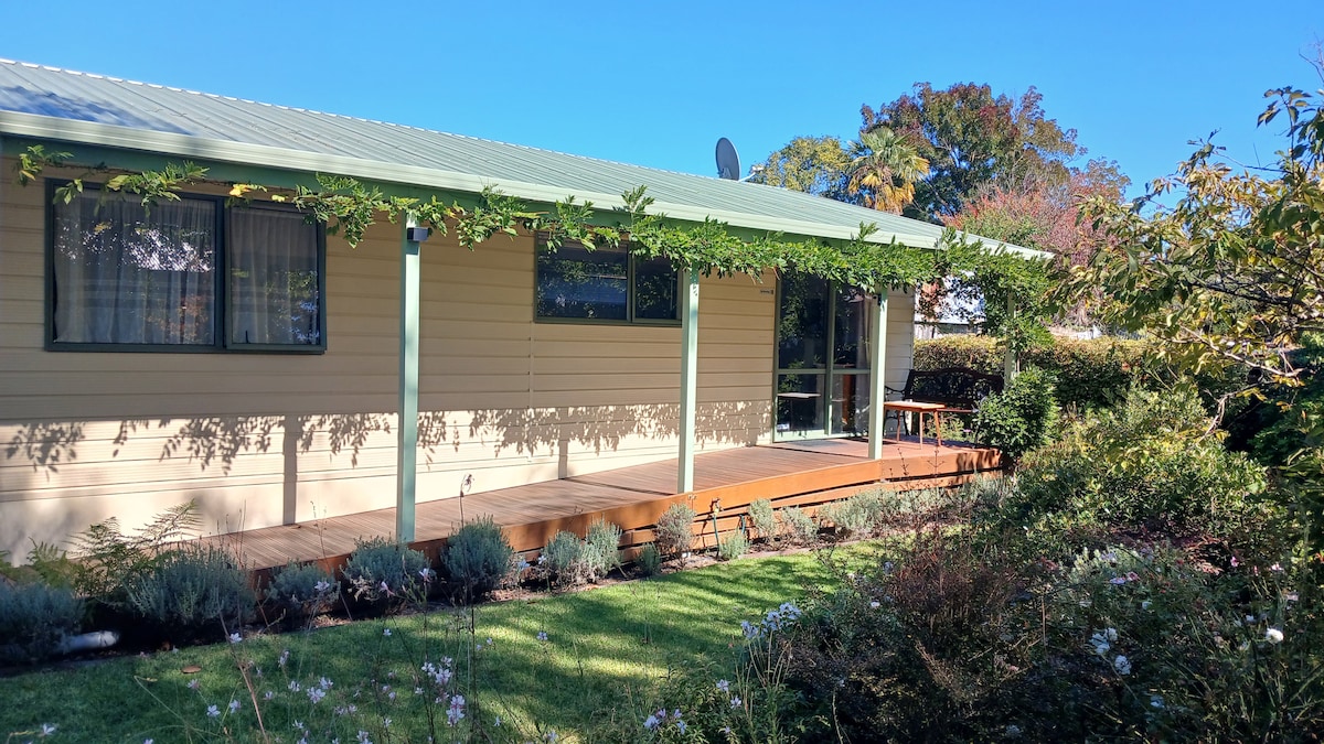 A single-story cottage is nestled within a lush garden, showcasing a covered porch that provides access to a large wooden deck. Windows with soft curtains allow natural light to flow in, while landscaped greenery surrounds the exterior, creating a peaceful setting.