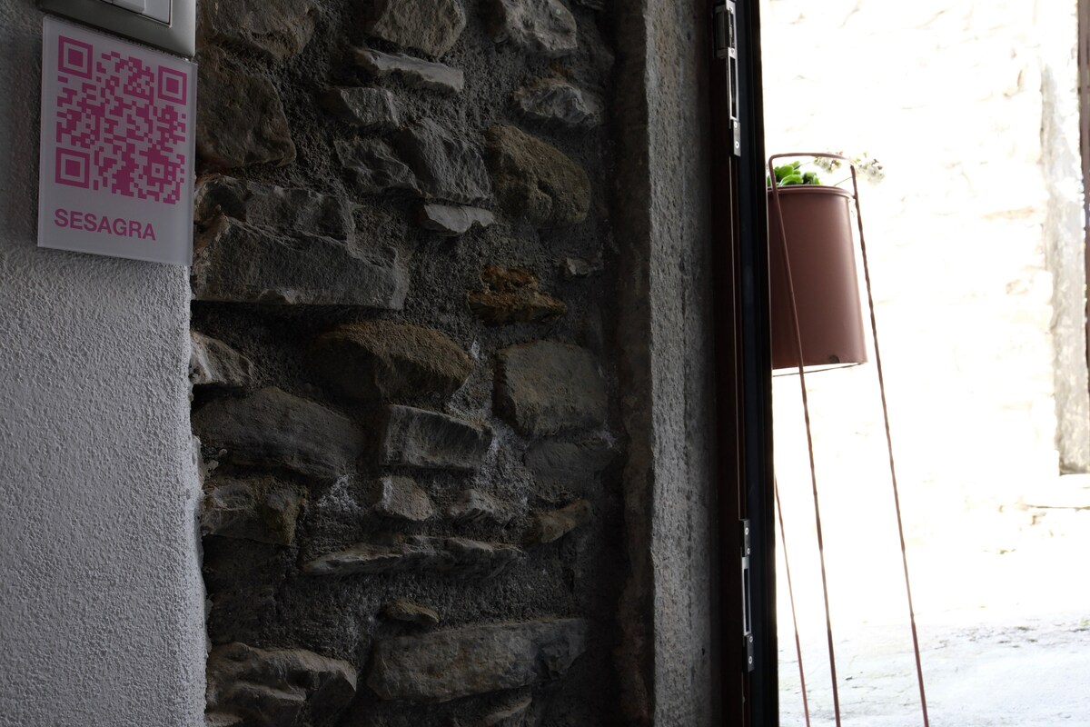 A rustic stone wall features a QR code sign near the entrance, providing visitors with digital access to information. Natural light streams in from an open door, illuminating a hanging planter that adds a touch of greenery to the stone texture.