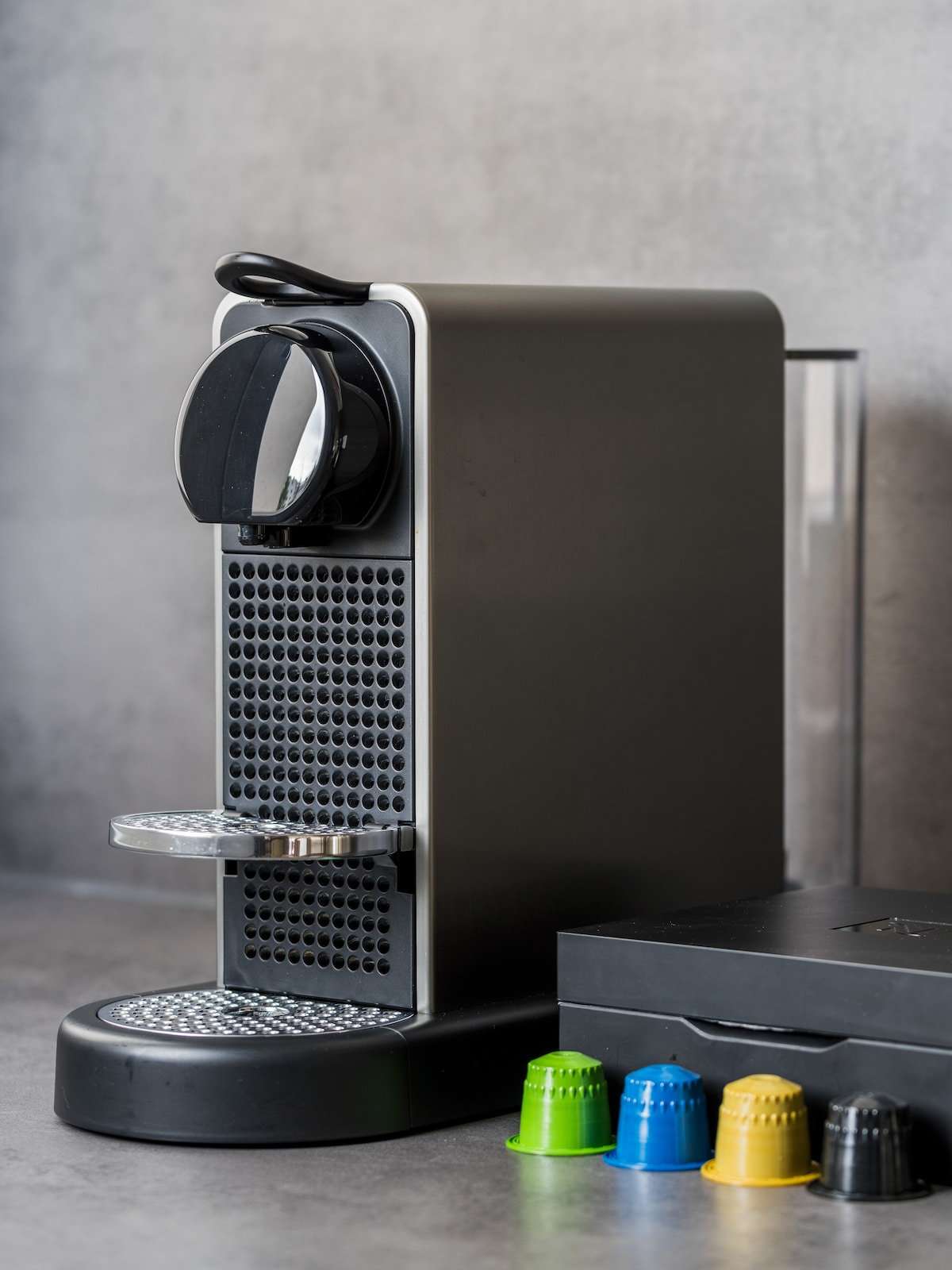 A modern coffee machine is placed on a sleek countertop, featuring a glossy finish and a circular control knob. A selection of colorful coffee capsules in green, yellow, and blue sits nearby, alongside a black storage box. The background consists of a minimalist gray surface.
