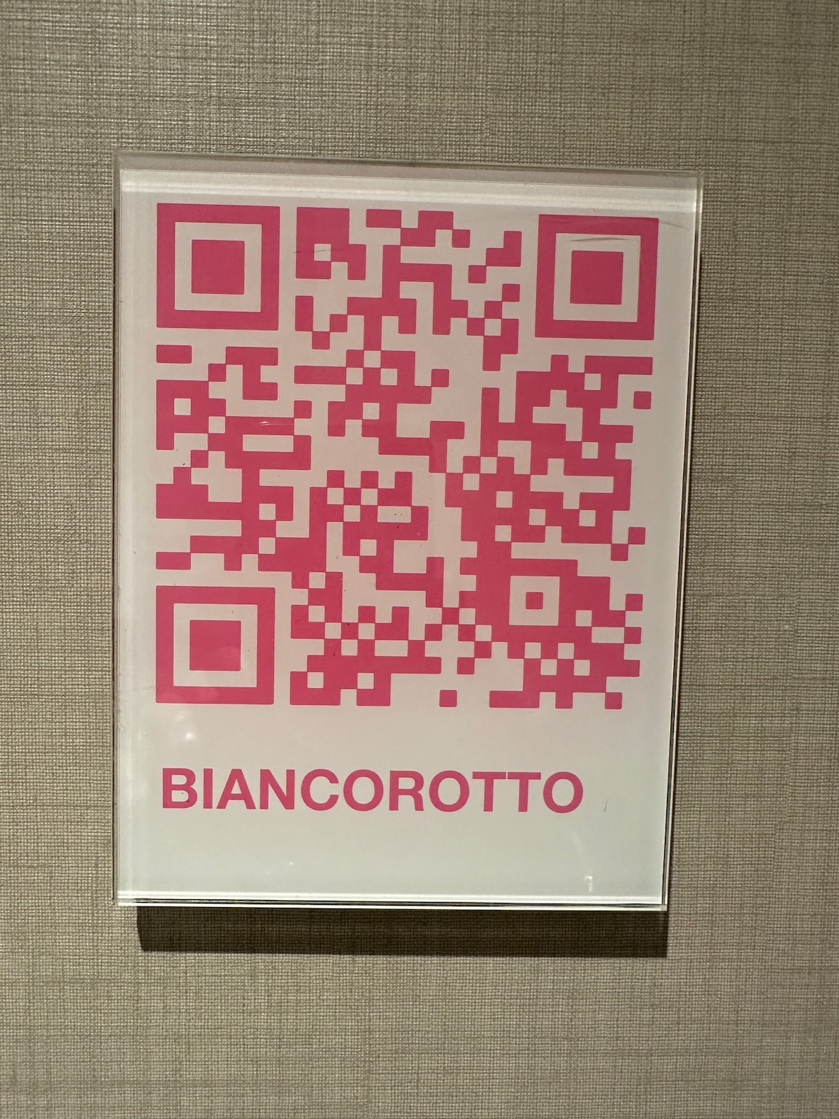 A framed sign is displayed on a textured wall, featuring a pink QR code centered above the name 'BIANCOROTTO' in bold pink lettering. The overall design is simple and modern, contributing to the room's contemporary aesthetic.