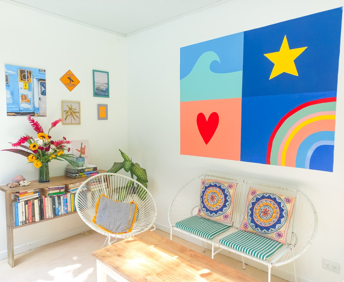 A bright living area features a large colorful wall art piece divided into four sections, displaying waves, a star, a heart, and a rainbow. Two white chairs with vibrant patterned cushions are placed around a wooden table, complemented by a bouquet of fresh flowers and framed artwork on the walls.