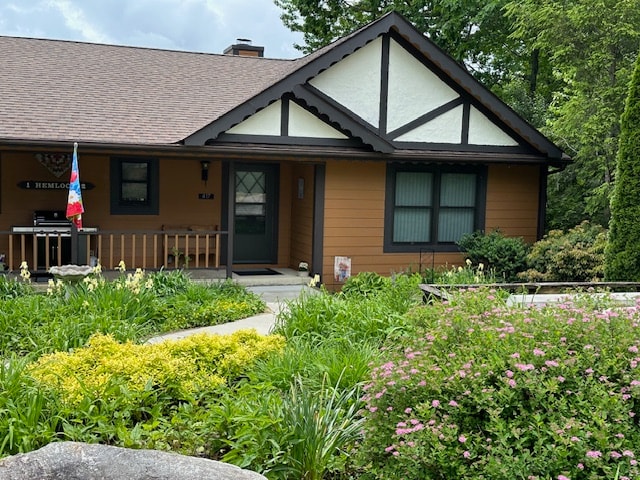 The cabin's exterior features a blend of wooden and stone elements, accented by flower beds filled with lush greenery and colorful blooms. A welcoming pathway leads to the entrance, with a porch displaying decorative touches and an inviting atmosphere. Soft natural light filters through the surrounding trees.