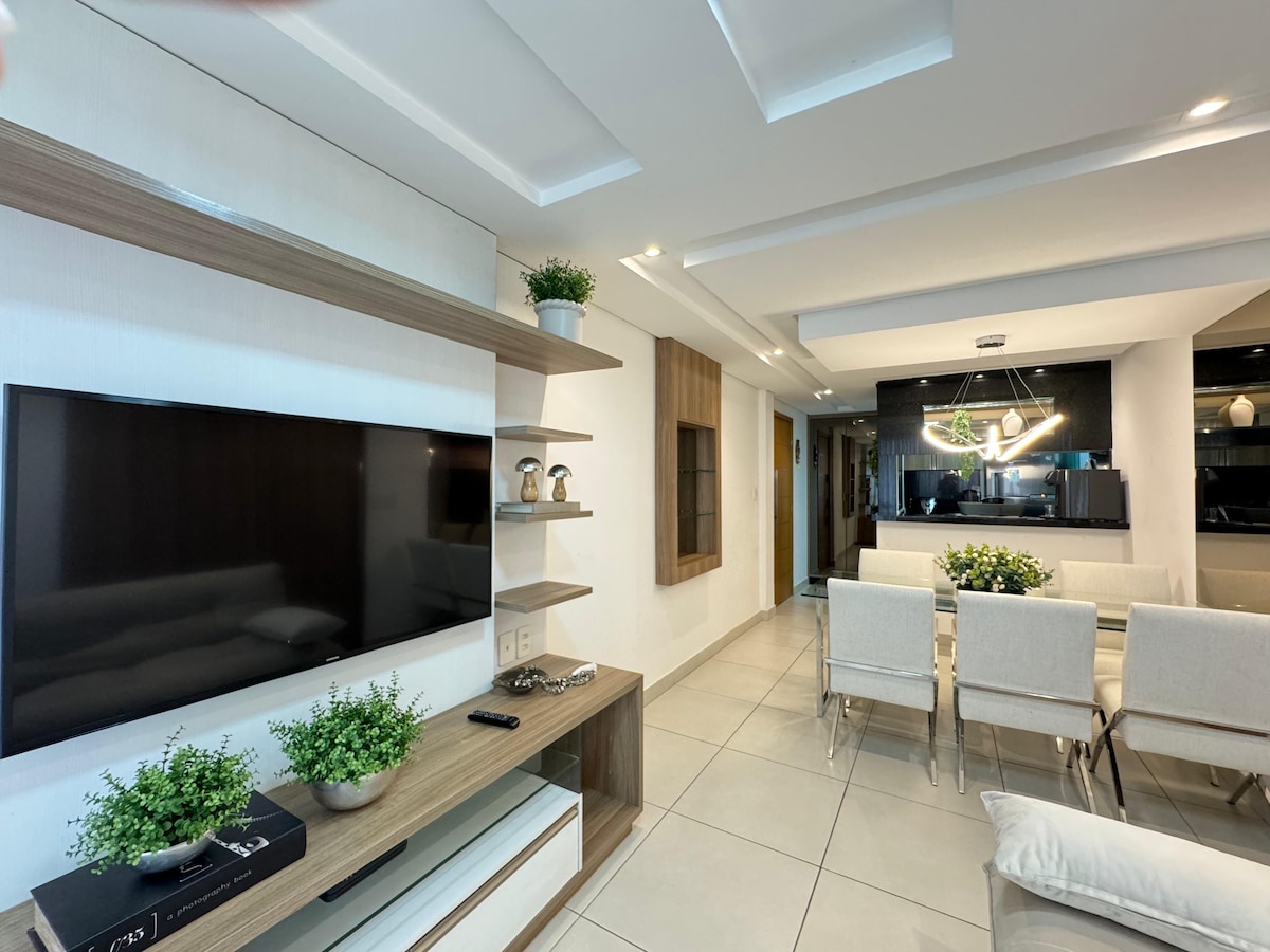 A modern living space is highlighted by a wall-mounted television and minimalist shelving, adorned with potted plants. A spacious dining area features a sleek table with upholstered chairs, complemented by neutral tones and ambient lighting, creating an inviting atmosphere.