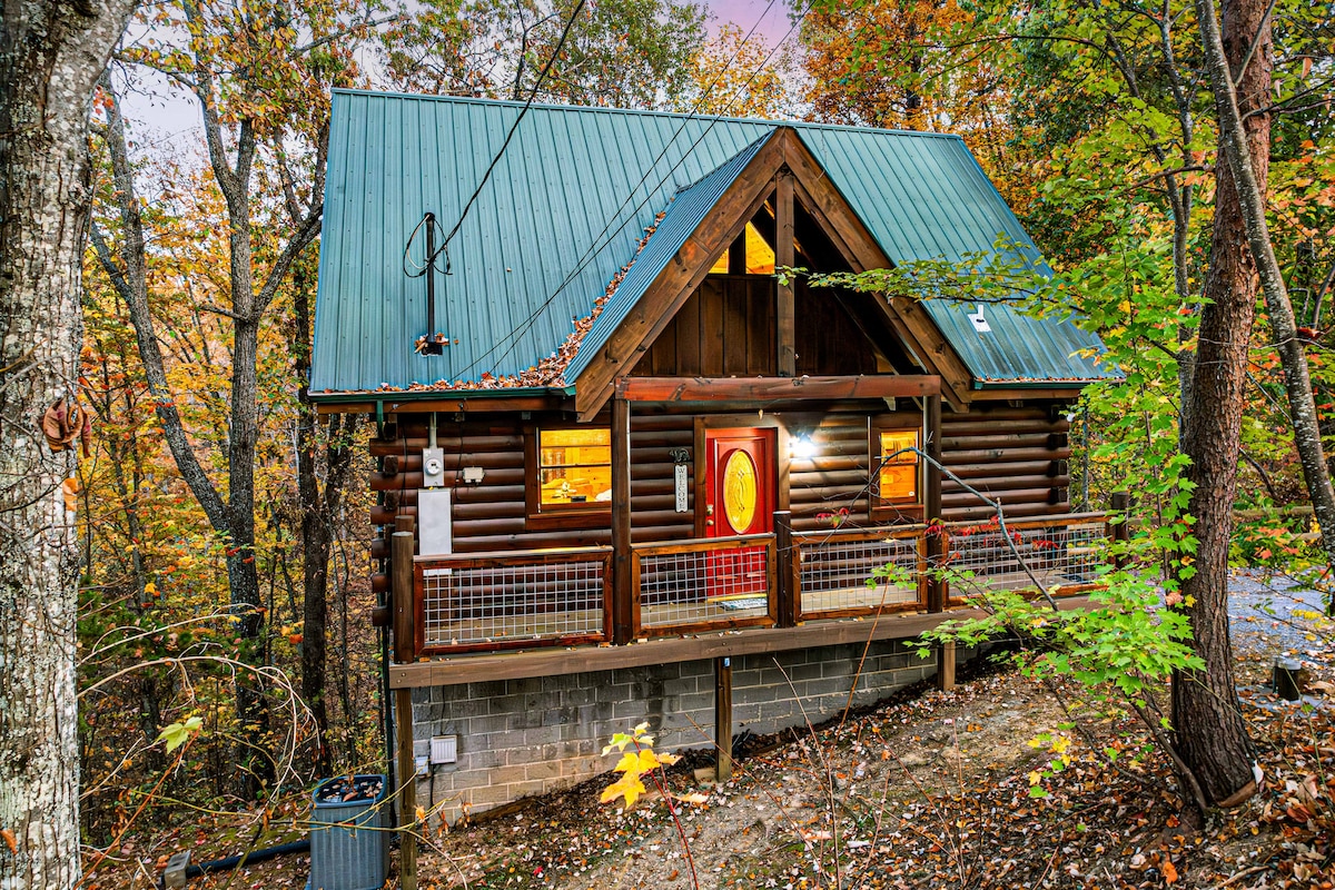 The cabin's exterior features a sturdy log construction with a green metal roof, surrounded by vibrant autumn trees. A spacious deck extends across the front, highlighted by a welcoming red door. Large windows provide views of the natural setting, emphasizing the cabin's connection to its surroundings.