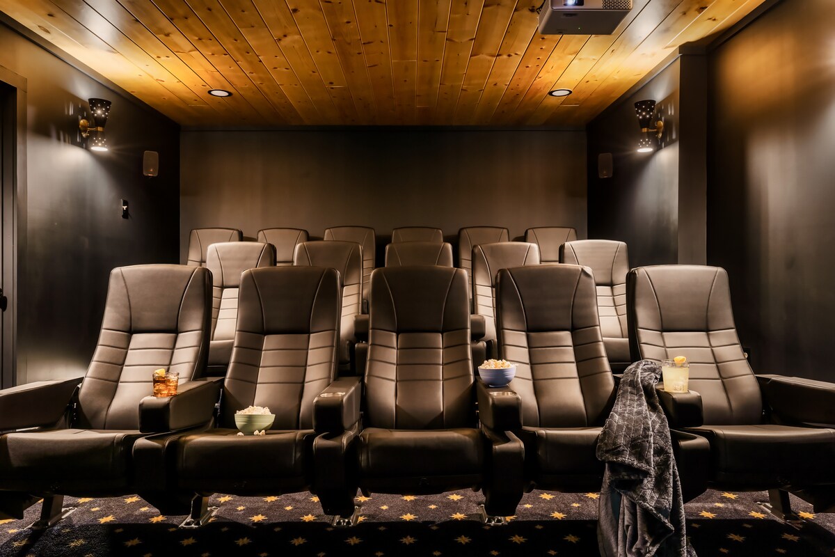 A private indoor movie theater features tiered seating for 16, with plush black reclining chairs arranged in rows. The space is dimly lit, with a rich wooden ceiling and a large projector screen at the front, designed for comfortable viewing.