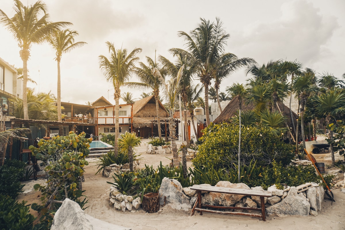 A lush garden area is surrounded by palm trees and tropical plants, creating a natural, serene atmosphere. Well-maintained paths lead to private structures, complemented by a mixture of thatched and modern rooftops. A bench made of stone rests in the foreground, enhancing the outdoor setting.