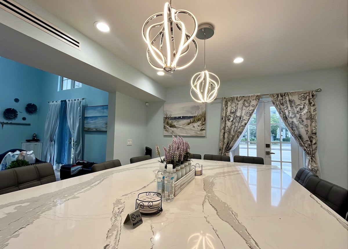 A large dining area features a spacious marble dining table surrounded by leather chairs. Unique light fixtures illuminate the space. Large glass doors open to the outside, complemented by decorative curtains and coastal artwork on the walls, enhancing the dining experience.