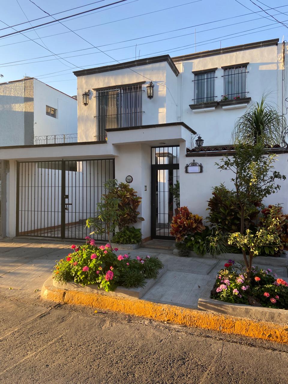 The exterior of the building is presented, featuring a modern two-story design with a gated entrance. Lush greenery and colorful flowering plants are arranged along the pathway, contributing to a welcoming atmosphere. Light reflects softly off the white walls under a clear sky.