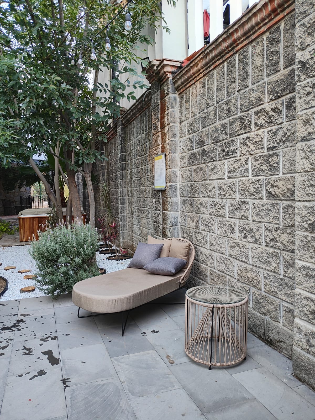 A cozy outdoor seating area features a curved lounge chair with soft cushions, positioned against a rustic stone wall. A small, round side table adds functionality, while greenery and pebbles enhance the natural ambiance of the space.