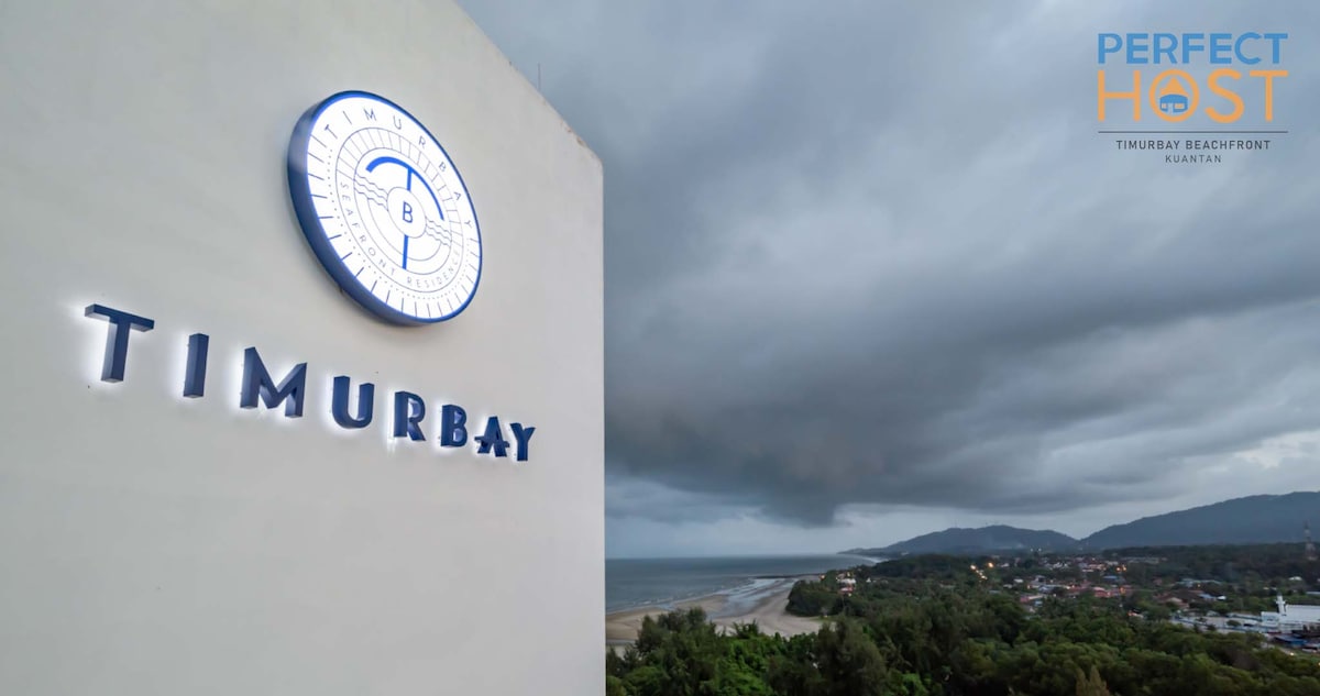 The exterior of Timurbay is prominently displayed against a dramatic sky, showcasing the beachfront's proximity. The logo is visible, highlighted by backlighting. Below, the beach meets the coastline, while the lush greenery and distant mountains create a natural backdrop.