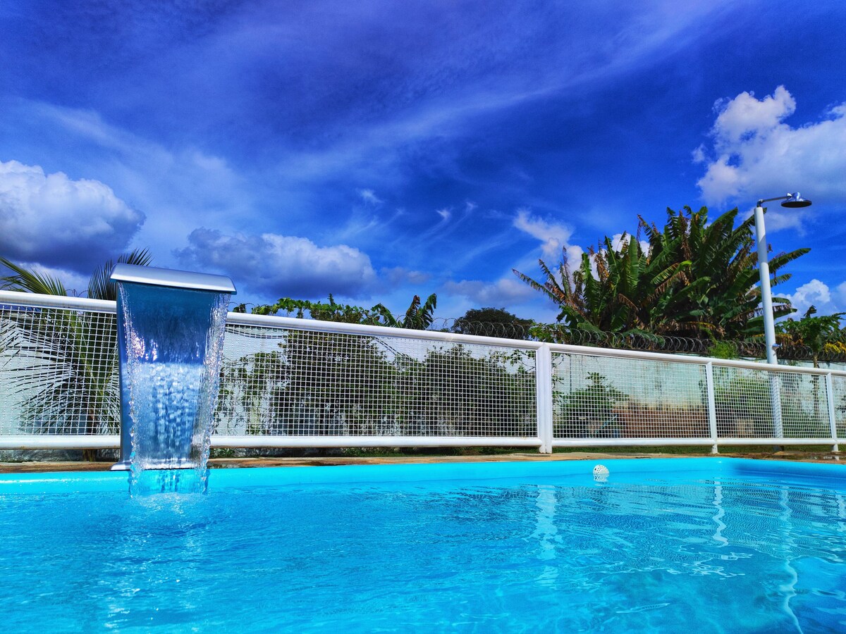 A clear, bright swimming pool is featured, with a gentle waterfall flowing into the water. Surrounding foliage includes green plants and trees under a vibrant blue sky, creating a serene environment for relaxation and leisure.