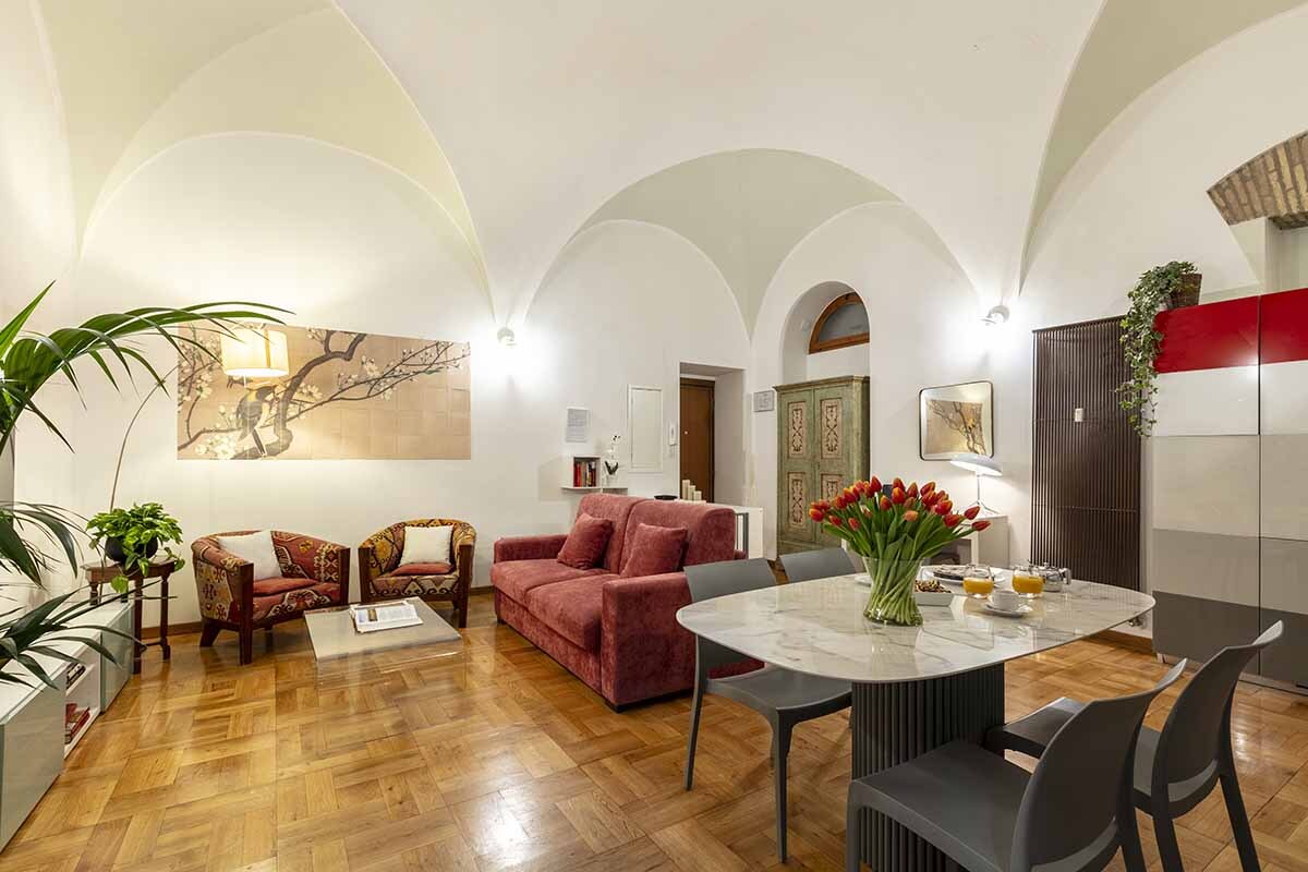 A spacious living area features high vaulted ceilings and a distinct architectural design. A plush red sofa is complemented by two patterned armchairs. A round dining table with stylish chairs is arranged nearby, adorned with fresh flowers and breakfast items for guests.