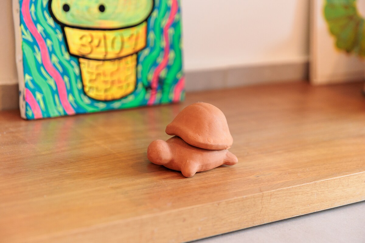 A sculpted terracotta turtle is positioned on a smooth wooden surface. Behind it, a colorful artwork adds vibrancy to the setting. The turtle's rounded shape and simple design highlight the creative decor elements within the space.