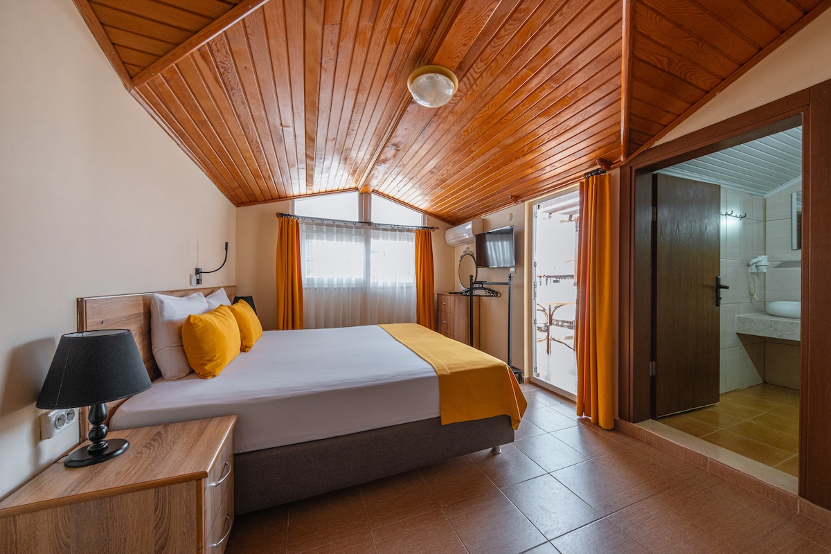 A bright sleeping area is showcased with a spacious bed centered against a warm wood-paneled ceiling. A desk with a lamp is positioned beside the bed. Natural light filters through a nearby window with sheer curtains, while an en-suite bathroom is visible through an open door.
