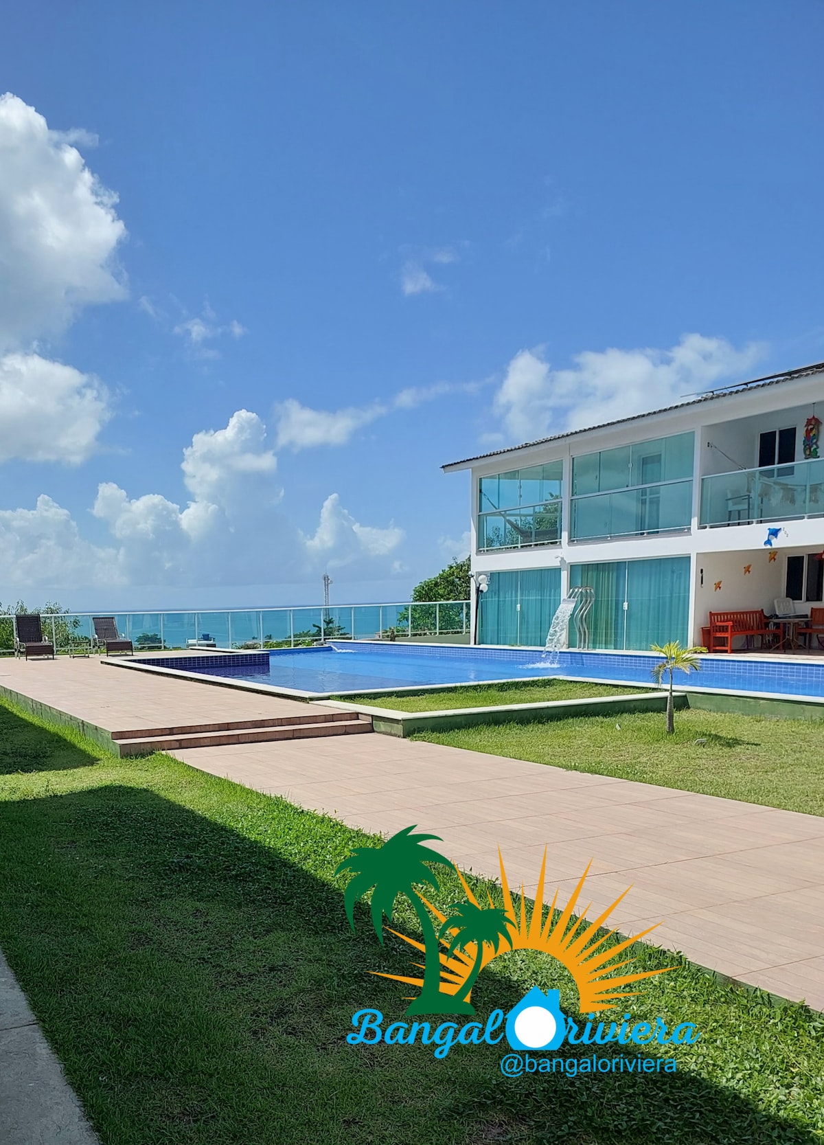 An outdoor view showcases a spacious swimming pool surrounded by lush green grass. The adjacent building features large windows that capture coastal views. A paved pathway leads from the pool area, providing access to the property and enhancing the open atmosphere.