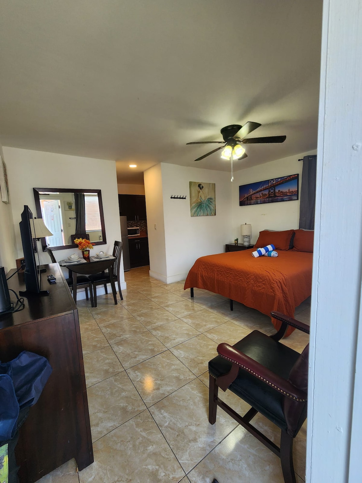 A spacious room is featured with an orange bedspread covering a large bed. A small dining table for two is positioned beside a sleek kitchen area. A wall-mounted television and a ceiling fan are also present, enhancing the functionality of the space.