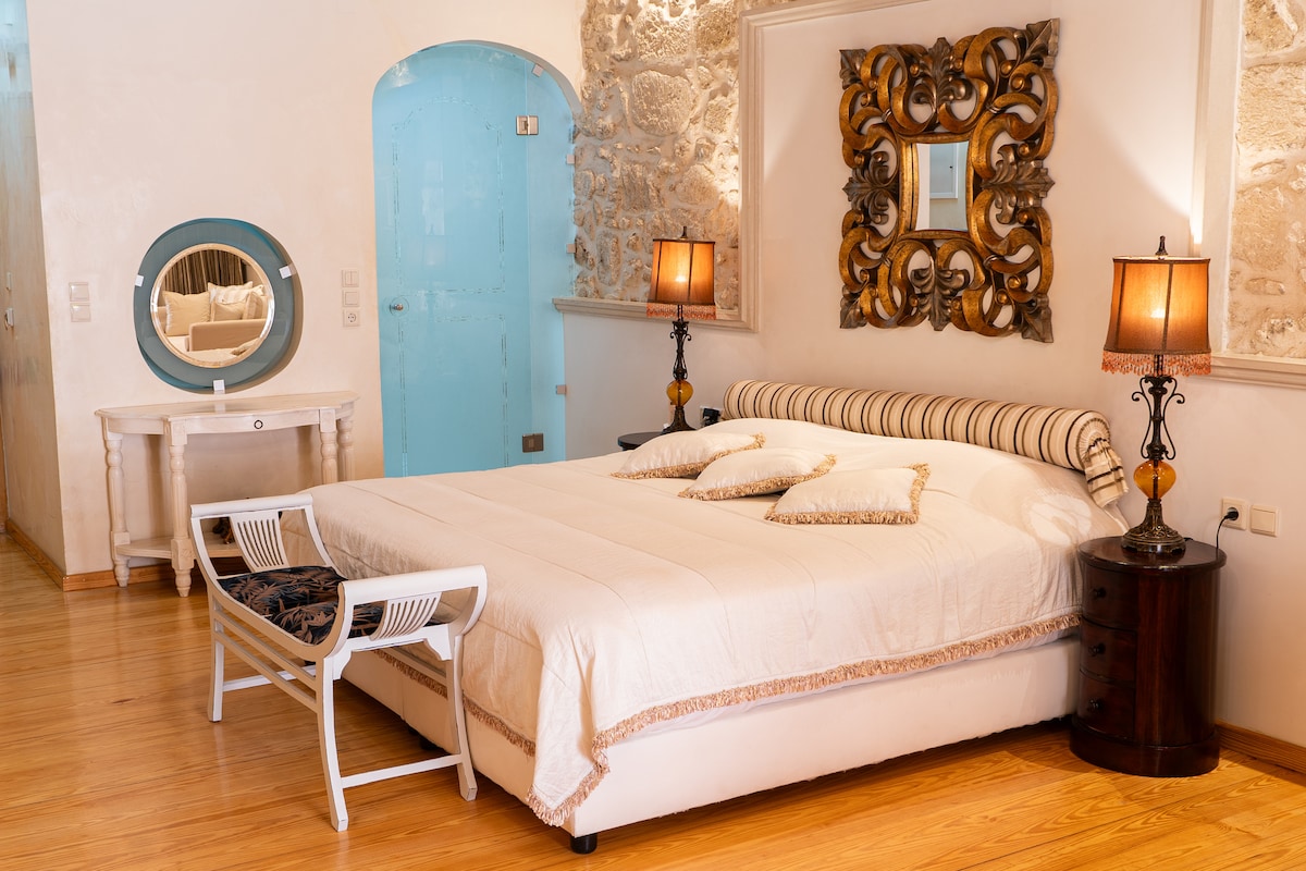 A spacious sleeping area features a large bed with decorative pillows and an elegant headboard. Two bedside tables with lamps flank the bed, while a mirrored vanity is positioned next to a light blue arched doorway. Exposed stone walls and wooden flooring add character to the room.