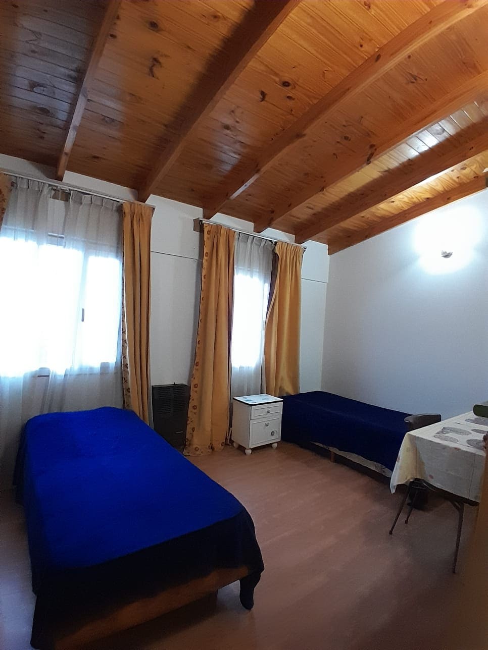 A serene bedroom features two twin beds dressed in blue bedspreads. Natural light enters through large windows adorned with sheer curtains. The wooden ceiling beams add warmth, while a small white nightstand and a table with chairs enhance the functional space.