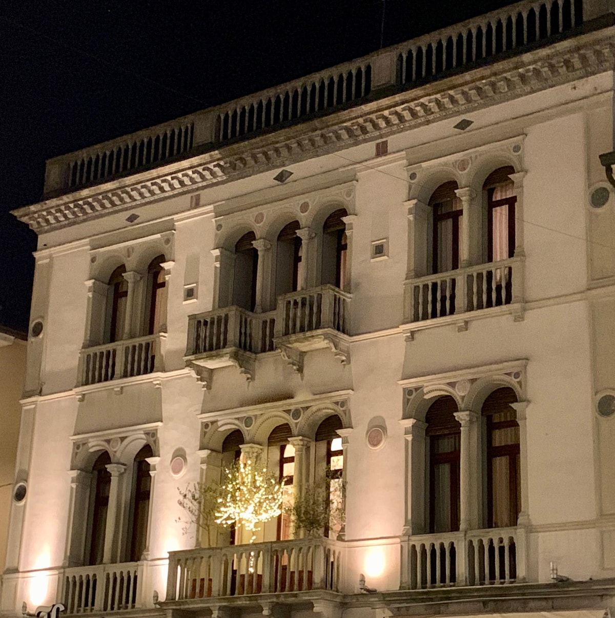 The historic building features elegant architecture with ornate arches and balconies. Soft lighting highlights the decorative details, creating a charming evening view. Potted plants adorn the balcony, adding a touch of greenery to the façade.