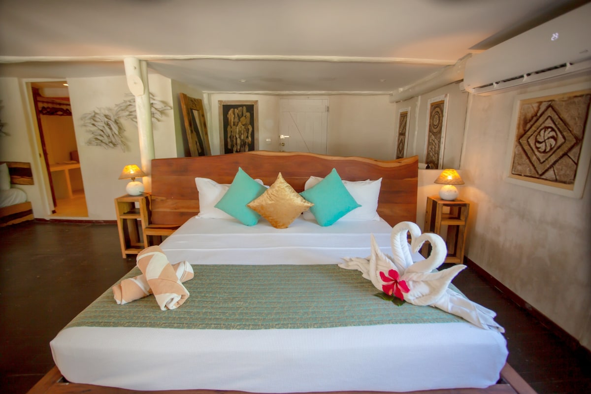 A spacious bedroom features a wooden bed adorned with colorful cushions in turquoise and gold tones. Decorative towel art resembling swans is present on the bed, alongside a vibrant flower accent. Soft lighting from bedside lamps enhances the calming ambiance.