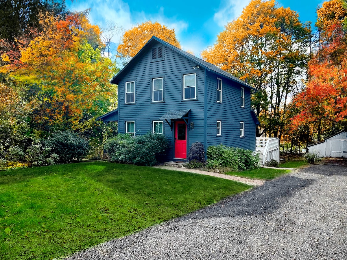 A charming two-story blue house features a vibrant red front door, surrounded by colorful autumn foliage and a spacious lawn. Gravel paths lead to the entrance, highlighting the inviting exterior and nearby greenery.