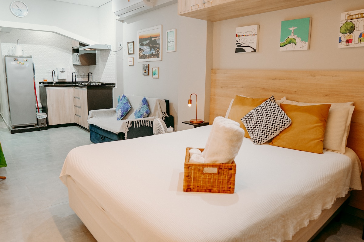 The loft features a comfortable queen-size bed adorned with decorative pillows. A cozy sofa bed is present next to the bed. The kitchen area includes modern appliances and a dining setup, with a decorative basket placed on the bed for convenience.