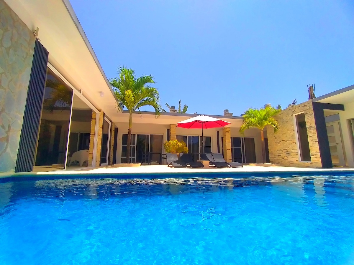 A modern villa features a clear blue pool surrounded by a patio area. Lounge chairs are positioned under a red and white umbrella, while tall palm trees provide a touch of greenery. Expansive sliding glass doors create a connection between indoor and outdoor spaces.