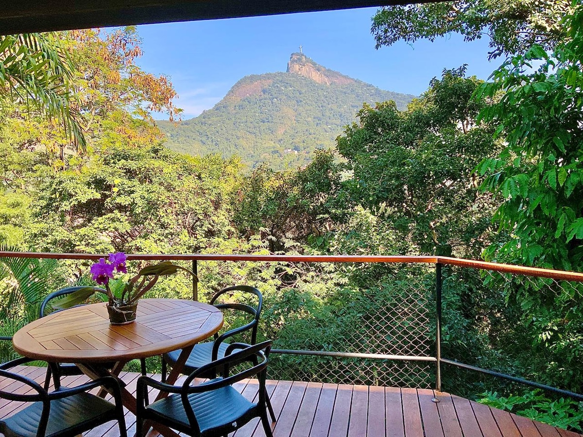 A private deck is highlighted, featuring a round wooden table surrounded by four black chairs. A potted purple orchid adds a touch of color. The view encompasses lush greenery and a mountain in the background, showcasing a clear blue sky.
