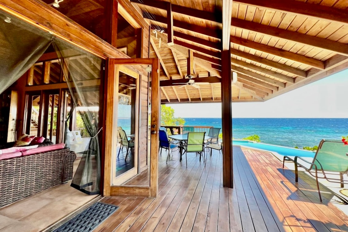 An open-layout living space features wood elements and large glass doors, connecting indoor and outdoor areas. A cozy seating area with plush cushions is visible alongside a dining set positioned for ocean views. The pool glimmers in the sunlight, enhancing the tranquil coastal ambiance.