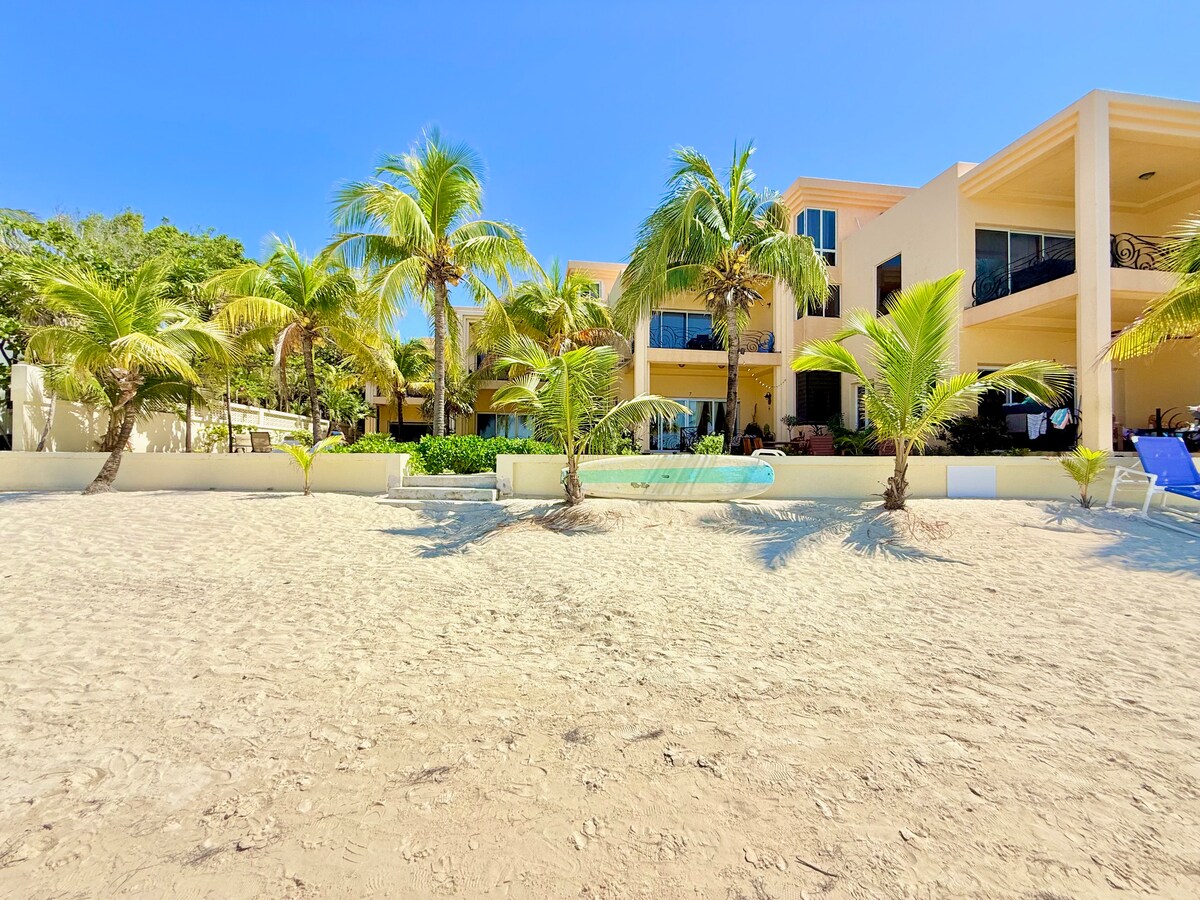 The exterior of the beachfront condominium is visible, showcasing palm trees and sandy landscaping. Sunloungers are positioned in the foreground, with the beach leading directly to the entrance. The background features the condo's structure with large windows and balconies, suggesting a relaxed atmosphere.
