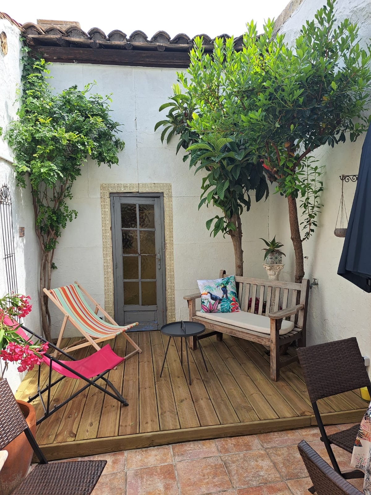 A private patio is surrounded by greenery, featuring two trees and colorful seating options. A wooden bench and a small table are positioned against a white wall, with bright chairs adding a touch of vibrancy. Potted plants enhance the serene environment.