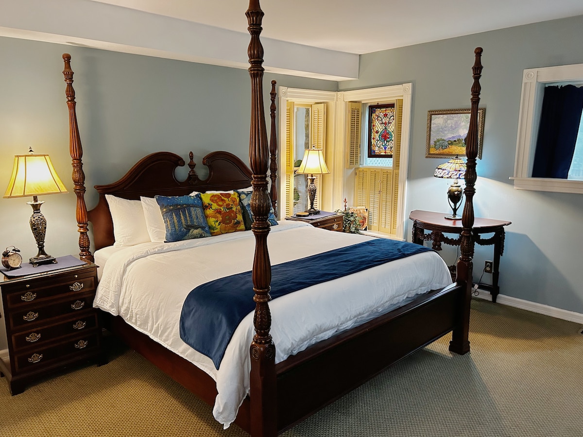 A spacious room features a four-poster king bed with a white comforter and decorative pillows. Soft lighting from two lamps complements the blue accent decor. A cozy sitting area is visible near a window, with a small table and a colorful wall art piece adding charm.